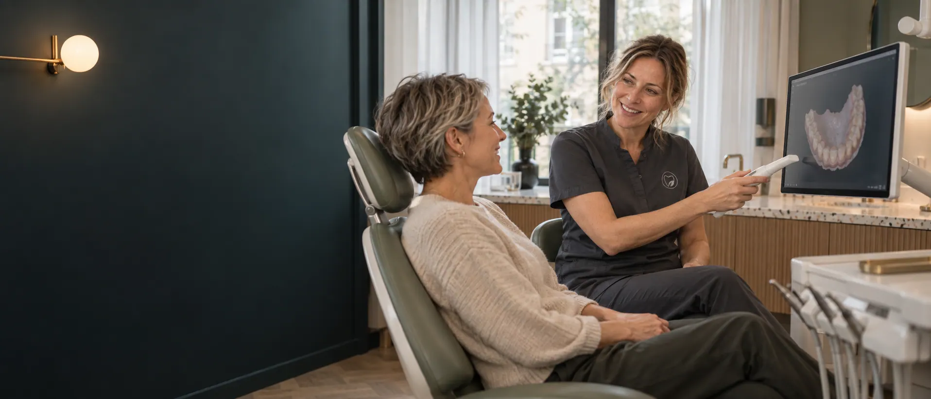 Dr. Johanna Weiss zeigt einer Patientin im Behandlungsstuhl den Intraoralscanner, auf dem Monitor ein 3D-Modell ihrer Zähne.